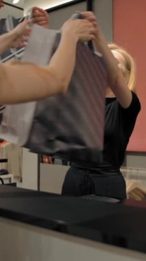 Genç bir adam ve kadının gülümseyen mağaza müdüründen kağıt poşet alıp kucaklaşırken ve birlikte yürürken görüntüsü. Dikey çekim.