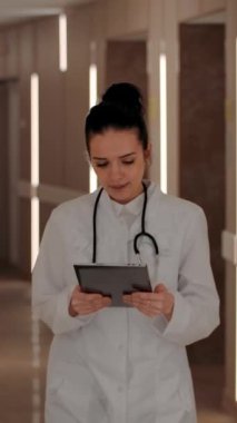 Neşeli kadın tıp doktoru klinik koridorunda yürüyor ve tabletten veri okuyor sonra durup kameraya gülümsüyor. Dikey çekim.
