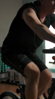 Siyah tişörtlü ve bandanalı orta yaşlı bir erkek sigara dolu bir odada durağan bir bisikletle tren kullanıyor. Spor salonunda eğitim aparatlarıyla yalnız bir sporcu. Yavaş çekim portre görünümü. Dikey çekim.