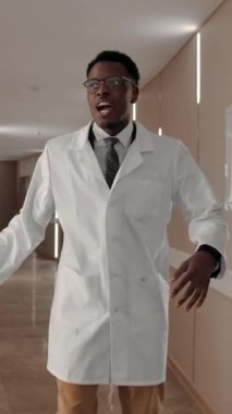 Heyecanlı Afrikalı Amerikalı tıp pratisyeni gülümsüyor ve dans ediyor, sonra kağıtları çarpıyor ve modern kliniğin koridorunda ağır çekimde hareket etmeye devam ediyor. Dikey çekim.