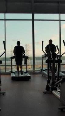 Güneşli bir günde boş bir spor salonunda büyük bir pencereye karşı koşu bandında koşan tanınmayan üç kişinin siluetleri. Sol kanat aşırı ağır çekimde. Dikey çekim.