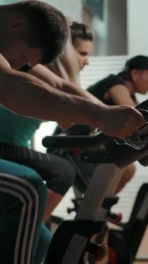 Üç sporcu yetişkin Kafkasyalı, iki erkek ve bir kadın bisiklet simülatörü. Modern bir spor salonunda sağlıklı bir yaşam tarzı. Yavaş çekim düzgün çekim. Dikey çekim.