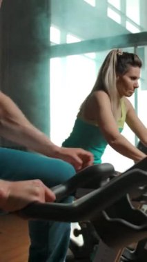 Sporcu iki arkadaşının yanında durağan bir bisiklete oturuyor ve pedal çevirmeye başlıyor. Modern bir spor salonunda sağlıklı bir yaşam tarzı. Dikey çekim.