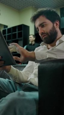 Genç sakallı, beyaz bir adam modern bir berber dükkanına girmek için sırasının gelmesini beklerken dijital ekranda sosyal güvenlik numarasını kontrol ediyor. Berber kız arka planda bir müşteriye saç stili yapıyor. Dikey çekim.