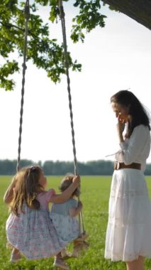 Genç bir anne, 1 ve 3 yaşlarındaki kızlarını yaşlı bir meşe ağacına bağlı ahşap bir salıncakta sallıyor. Sevgi dolu bir anneyle tasasız mutlu çocukluk kavramı. Gündoğumu turuncu ışığı. Dikey çekim.