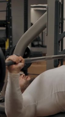 Güçlü erkek ağırlık kaldırırken halterle bankta yatıyor. Kas sporları eğitmeni modern bir spor salonunda güç antrenmanı yapıyor. Yavaş çekim dikey çekim.