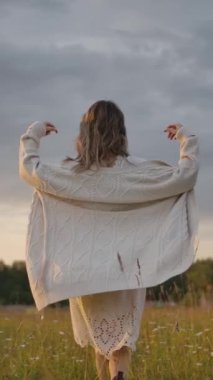 Cazibeli bayan elbisenin üzerine uzun örülü hırka giyip kırsal bölgedeki bulutlu gökyüzüne karşı çimen tarlasında dönüyor. Dikey çekim.