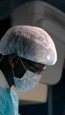 Siyahi doktorun kameraya dönüp, ameliyatın başarıyla bitmesinin ardından maskesini çıkarıp kameraya gülümsüyor. Dikey çekim.