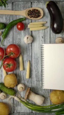 Ahşap tezgahın üzerine sebze malzemeleri ve baharatlarla atılmış beyaz boş sayfalı yemek tarifi kitabının üst görüntüsü. Makbuzunu boş bir kağıda koymak kolay. Dikey çekim.