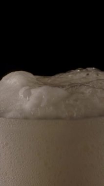 Koyu bardaki yumuşak köpüklü taze soğuk bira bardağına yakınlaştıran kamera. Dikey çekim.