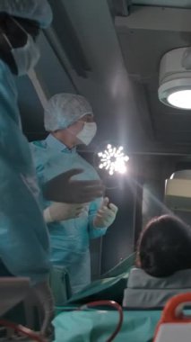 Esmer tenli ve beyaz cerrahlar ameliyathanede ameliyat hakkında konuşuyorlar. Ameliyat masasının altında yavaş çekim geniş kamera hareketi. Dikey çekim.
