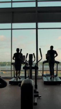 Güneşli bir günde boş bir spor salonunda büyük bir pencereye karşı koşu bandında tanınmayan üç kişi koşuyor. Sağdan sola yumuşak, yavaş çekim. Dikey çekim.