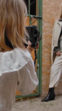 Arka planda oturan bir kadın, zarif bir kadın modelin çiftlikteki ahır duvarının yanında saçlarını çekerken fotoğraflarını çekiyor. Dikey çekim.