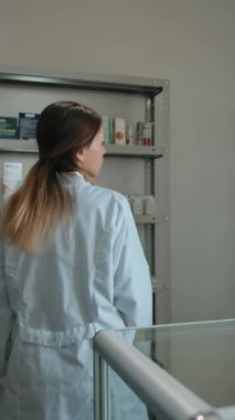 Eczanenin iç tarafındaki eczacı kızı vurdular. Beyaz bornozlu güzel bir kadın eczacı kameraya bakar ve gülümser. Raflardaki malları kontrol ediyorum. Dikey çekim.