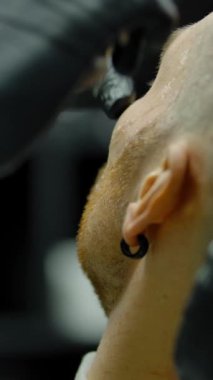 Berber dükkanında saçları boyalı bir erkek müşterinin yanağını tıraş etmek için düz jilet kullanan eldivenli tanınmaz bir berber. Dikey çekim.