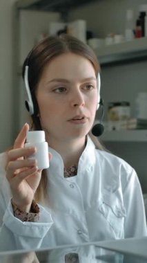 Kulaklık takmış genç bir kadının portresi, hapları kavanozda müşteriye gösteriyor, eczanede. Kafkasyalı kadın eczacı internetten potansiyel bir eczane müşterisiyle modern tıptan bahsediyor. Dikey