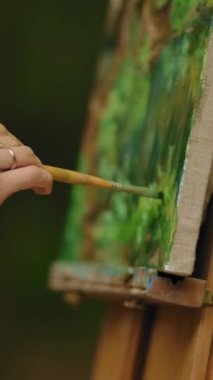 Bir kadın eli, bir fırçayla peyzaj resmi çiziyor. Doğada özgür bir sanatçı tarafından yapılan son dokunuşlar. Açık havada manzara çizmek en sevdiği hobidir. Dikey çekim.