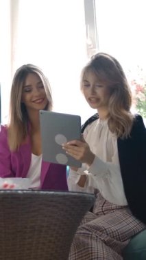 Moda kıyafetleri içinde, gülümseyen ve giysi seçmek için tablet kullanan iyimser genç kadınlar elbise mağazasında kanepede otururken. Dikey çekim.
