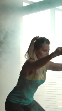 Kız spor salonundaki yarımkürede çömeliyor. Kalçalar üzerinde ustalık isteyen bir egzersiz. Duman dolu spor salonu, büyük pencereler, stüdyo ışığı, güzel bir kadın vücudu silueti. Dikey çekim.