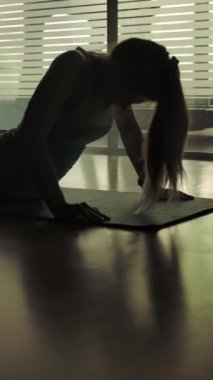 Spor salonunda spor halısı üzerinde yoga yapan seksi bir kadının silüeti. Göğsünü açıyorum. Pencereden akşam güneşinin ışığı. Dikey çekim.