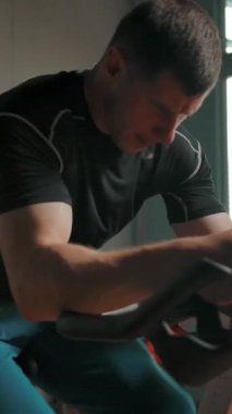 Spor salonunda bisiklete binen etkin bir erkek sporcu. Güç konseptin ayaklarına yüklenir. Bu sıkı bir egzersiz. Ağır çekim. Dikey çekim.