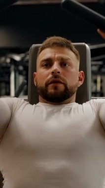 Güçlü kaslı adam egzersiz makinesinde pazı egzersizi yapıyor. Modern bir spor salonu, arka planda çeşitli spor aletleri. Dikey çekim.