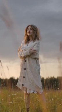 Örgü örmüş, uzun hırka giyen, gözlerini kapayan, kollarını kucaklayan, sonra da bulutlu gün batımına karşı çimenli çayırlarda yürüyen zarif bir bayan. Dikey çekim.