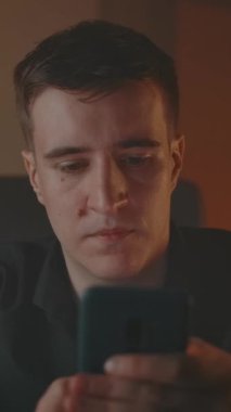 Dumanlı, rahat bir akşam odasında akıllı telefondan mesaj yazan bir adam. Beyaz tenli yakışıklı erkek sosyal ağlarda sörf yapıyor. Evin içinde, yakın planda. Dikey çekim.