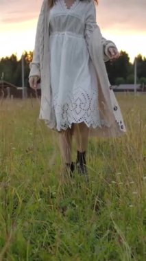 Beyaz elbiseli ve uzun ceketli genç bir kadının çimenlerin üzerinde yürürken ve rüya gibi bir bakışla yukarı bakarken, sonra gün batımının bulutlu gökyüzüne gülümserken görüntüsü. Dikey çekim.