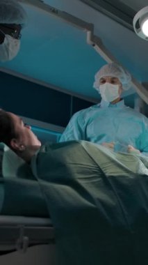Koyu tenli bir cerrah ve ameliyathanedeki meslektaşı karmaşık bir ameliyat için kadın bir hastayla birlikte bir kanepeyi röntgen cihazının altına taşıdılar. Dikey çekim.
