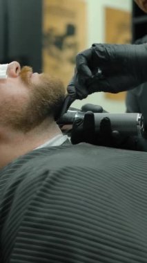 Berber dükkanında çalışırken gözlerinde doku olan erkek müşterinin sakalını taramak için elektrikli tıraş makinesi ve tarak kullanan yetişkin bir adamın etrafından dolaşın. Dikey çekim.