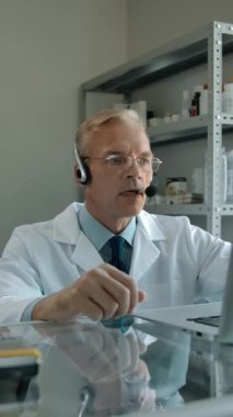 Eczacı, beyaz kağıt pakette müşteriler için online sipariş hazırlıyor. Beyaz erkek patron, kulaklık ve dizüstü bilgisayar kullanarak müşteriyle konuşuyor. Güzel bayan çalışanı teslimata hazır.