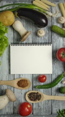 Tanınmayan şef tarifi incelerken havuç yiyor. Bir sürü farklı sebze, domates, biber, patlıcan, patates ve mantar ve ahşap bir masada bir tarif defteri. Dikey çekim.