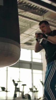 Boxer Gölge Boksu. Boksör yumruk atıyor, tekmeliyor ve kum torbasına tekme atıyor. Alt manzara. Modern fitness ekipmanları ve arka planda büyük pencereler. Dikey çekim.