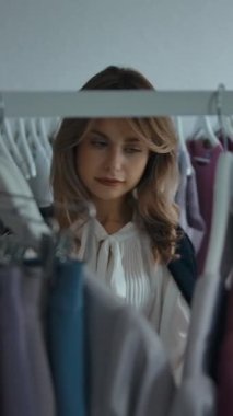 Akıllı telefonlu genç bir kadın raftan yeni elbiseler seçiyor ve hafta sonu alışverişi sırasında şık kıyafetlerle elbise asıyor. Dikey çekim.