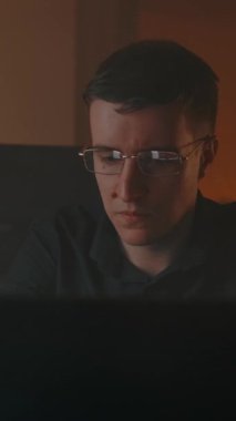 Gözlüklü, zeki, beyaz erkek. Ev içi rahat bir evde dizüstü bilgisayarda çalışıyor. Masanın üzerinde sanal gerçeklik kaskı var. Gece havası. Dikey çekim.