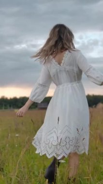 Beyaz elbiseli heyecanlı bir kadının, kara kedinin yanındaki yeşil çayırda, bulutlu gün batımına karşı gülümseyen ve dönen fotoğrafını çekiyorum. Dikey çekim.