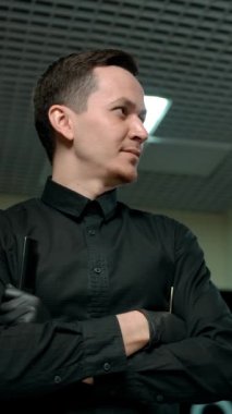 Çağdaş kolları çapraz olan koyu renk elbiseli kendine güvenen erkek berber modern bir berber dükkanının önünde duruyor. Önce sağa, sonra da kameraya bakıyor. Yavaş çekim portresi. Dikey çekim.