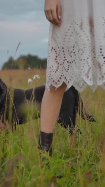 Bulutlu bir akşamda tarlada, beyaz elbiseli bir kadının yanında yeşil çimenlerde yürüyen siyah köpeğin görüntüsü. Dikey çekim.