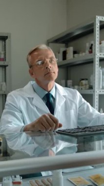 Kadın beyaz pakette siparişini ödemek için eczaneye geldi. Beyaz cüppeli ve gözlüklü beyaz erkek satıcı paketi veriyor ve kredi kartıyla ödeme yapmaya hazırlanıyor.