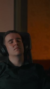 Kafkasyalı bir adam müzik dinlerken rahat bir evde koltukta dinleniyor. Büyük kulaklıklar, meditasyon için melodik müzik ve dizüstü bilgisayarın arkasında dinlenme. Gece dikey görünüyor.