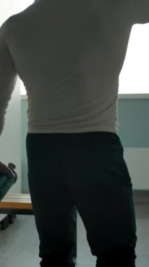 Beyaz erkek, güzel bir figürle soyunma odasına giriyor ve dinlenmek ve su içmek için bir bankta oturuyor. Kas ve kardiyo egzersizlerinden dinleniyorum. Dikey çekim.