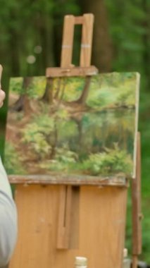 Kadın ressam, resminin resmini akıllı telefondan çekiyor. Esmer kadın, ormanda açık havada manzara resmediyor ve fotoğraf çekiyor. Arkadan bak. Yavaş parola. Dikey çekim.