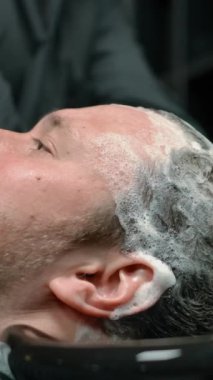 Eldiven giymiş, musluk takan ve berber dükkanında çalışırken sakallı adamın temiz saçlarından sabun köpüğünü yavaş çekimde lavaboya atan tanınmamış bir işçinin manzarası. Dikey çekim.