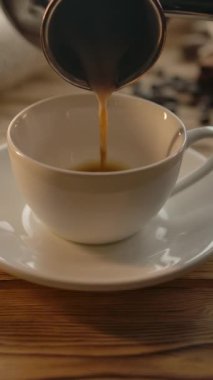 Cezve 'den gelen kahveyi ahşap bir masanın üzerindeki fincana döküyorum. Kese kağıdı, yulaf ciğeri ve kahve çekirdekleri. Kahvaltı için sıcak bir ortam. Ağır çekim. Dikey çekim.