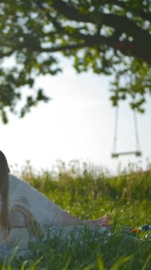 Genç bir anne ve iki küçük bebeği yalnız yaşlı bir meşe ağacının yakınındaki bir açıklıkta piknik yaptılar. Annem kızlarına sevgiyle bakıyor. O, o kaygısız oyuncakları oynarken izlemekten mutlu. Ağır çekim. Dikey çekim