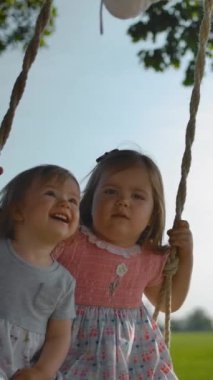 İki küçük kız salıncakta sallanıyor. Annem iki küçük kız kardeş, 1 ve 3 yaşında, halat saplı ahşap bir salıncak üzerinde sallar. İki çocuğun yüzleri yakın çekim. Mutluluk ve komik çığlıklar. Yavaş çekim