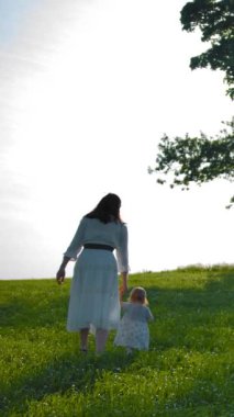 Küçük bir buçuk yaşındaki kızı ile anne bir salıncak ile ağaca doğru alan boyunca yürümek. En büyük kızı sallara gitmek için çok ilerigitti. Güneşli sıcak yaz günü gün batımı. Geniş atış. Geri görünüm