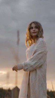 Romantik büyüleyici kadın yürüyor ve gülümsüyor, sonra uzun ceketini düzeltiyor ve bulutlu gün batımına karşı çimenli çayırda dönüyor. Dikey çekim.