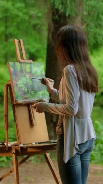 Manzara çizen genç bir sanatçının arka görüntüsü. Çekici, ince esmer kız dışarıda parkta bir resim çiziyor. Sanatta profesyonel. Bulutlu ışıklandırma dikey çekim.